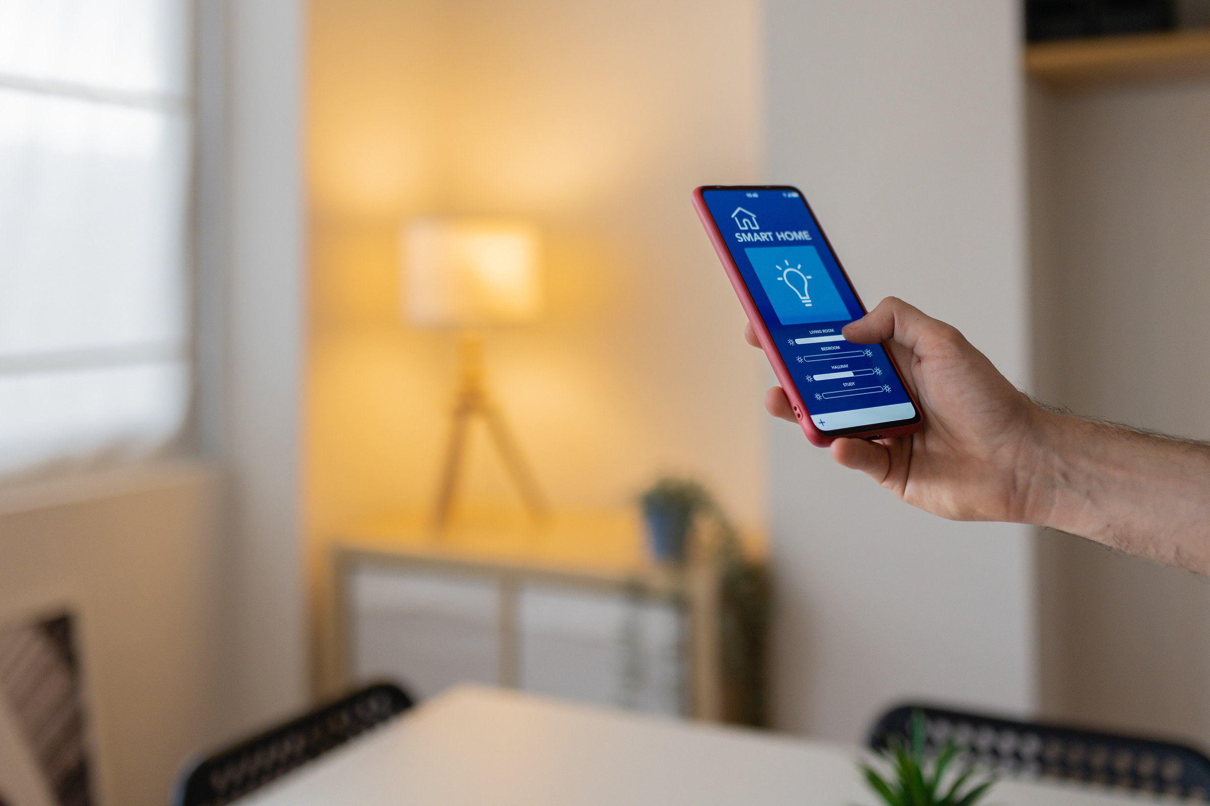 Dłoń ze smartfonem, na którym włączona jest aplikacja Smart home, na tle nowoczesnego, jasnego wnętrza.
