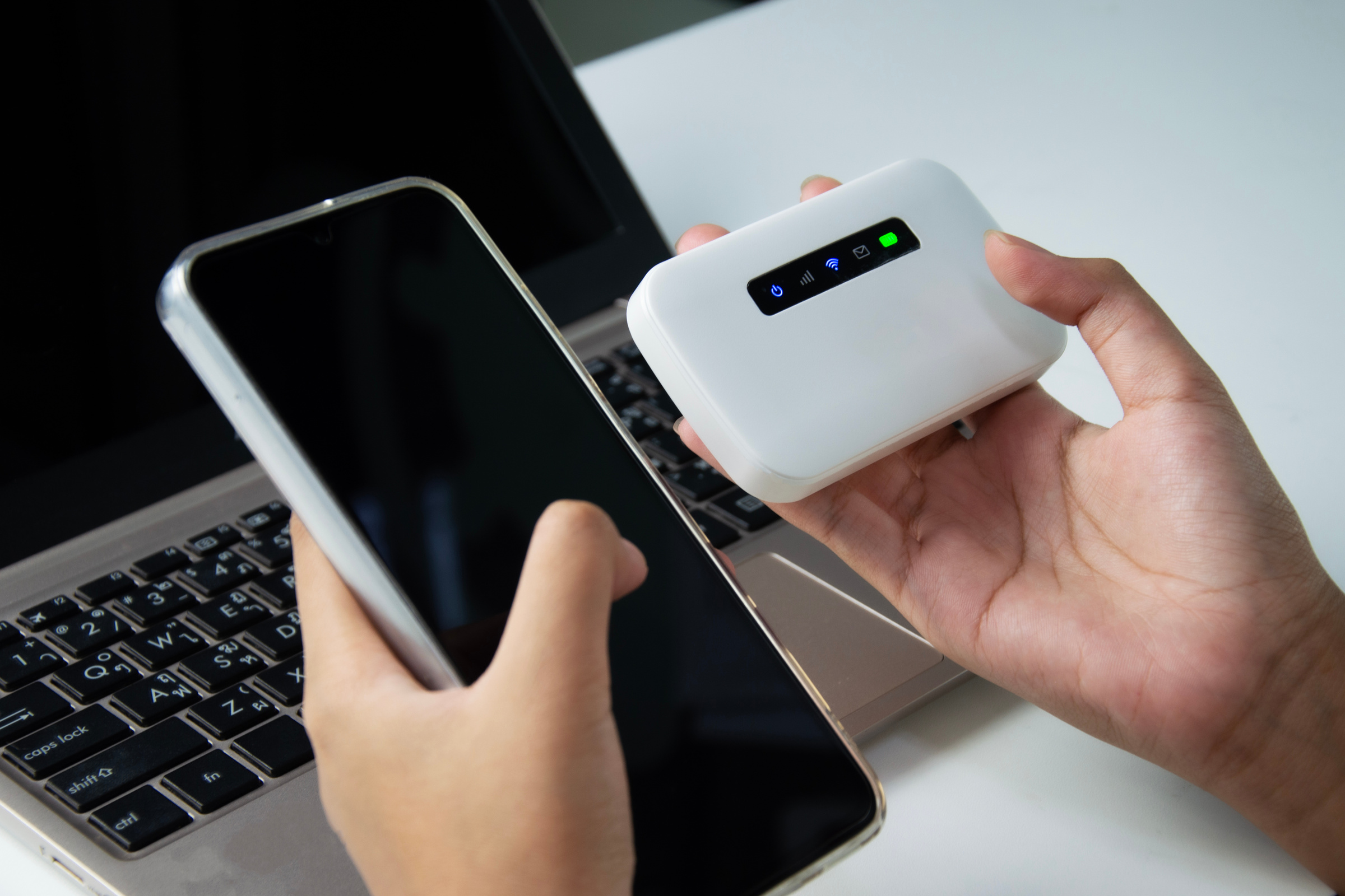 Smartfon i router mobilny trzymane obok siebie w dłoniach na tle laptopa.