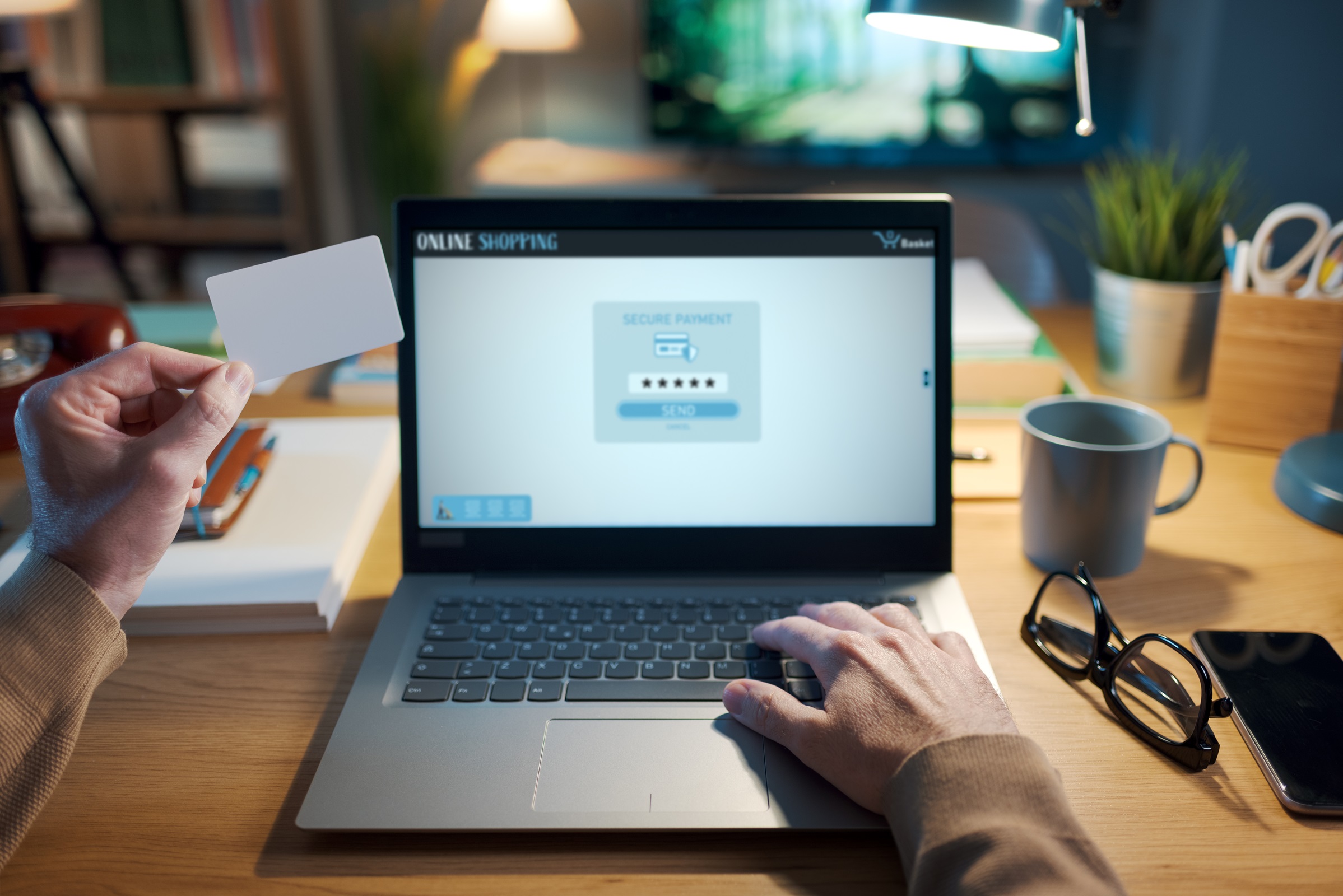 Laptop na drewnianym biurku z widoczną dłonią na klawiaturze wpisującą hasło, a w drugiej z kartą.