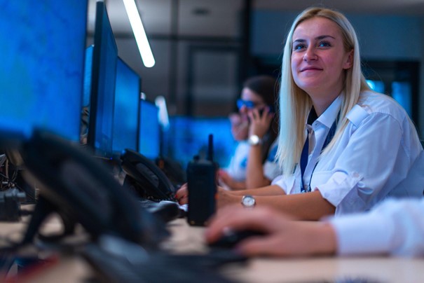 Uśmiechnięta kobieta w koszuli znajdująca się przy biurku wśród innych pracujących osób.