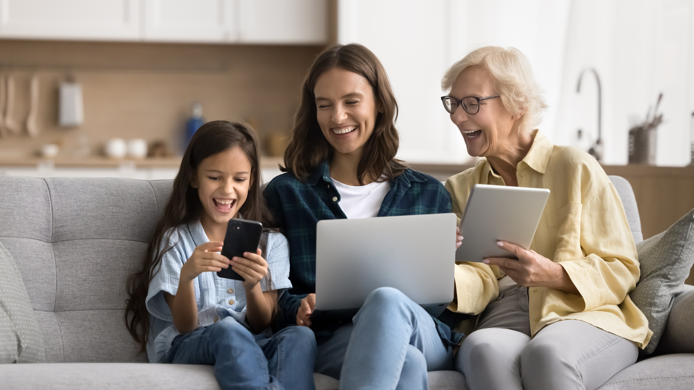 Uśmiechnięta mama, babcia i córka siedzące na kanapie z laptopem, tabletem i smartfonem.