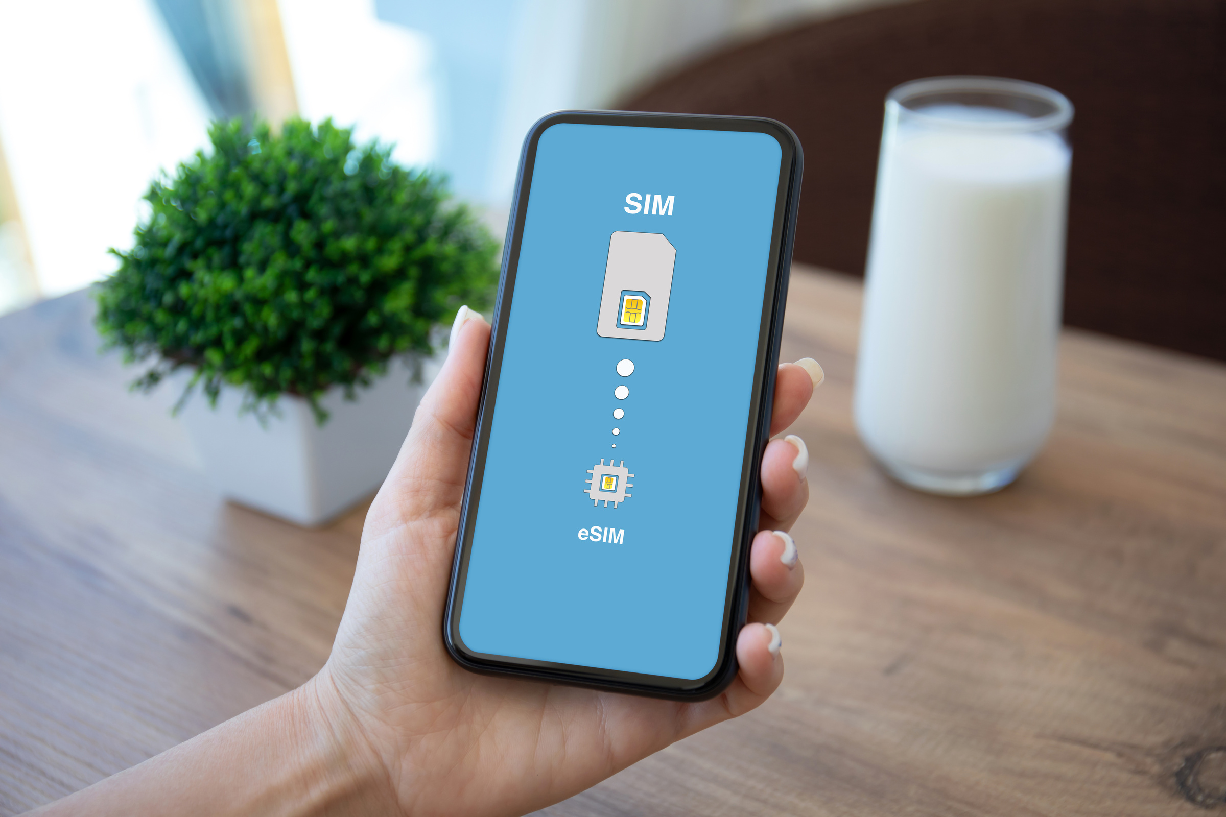 Smartfon w dłoni na tle drewnianego stołu z ikonkami na ekranie SIM przechodzący w eSIM
