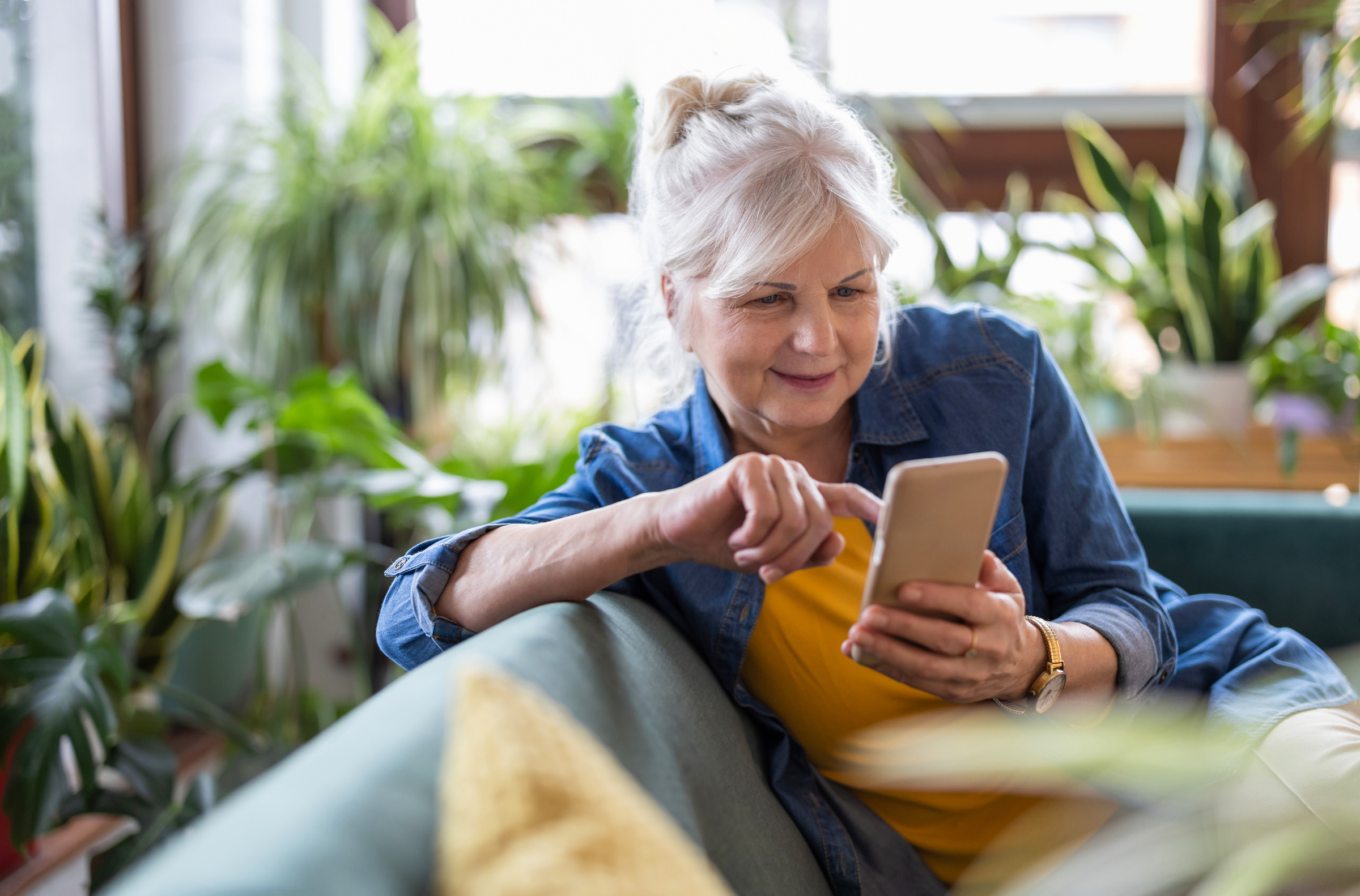 Uśmiechnięta seniorka siedząca na kanapie i obsługująca smartfon