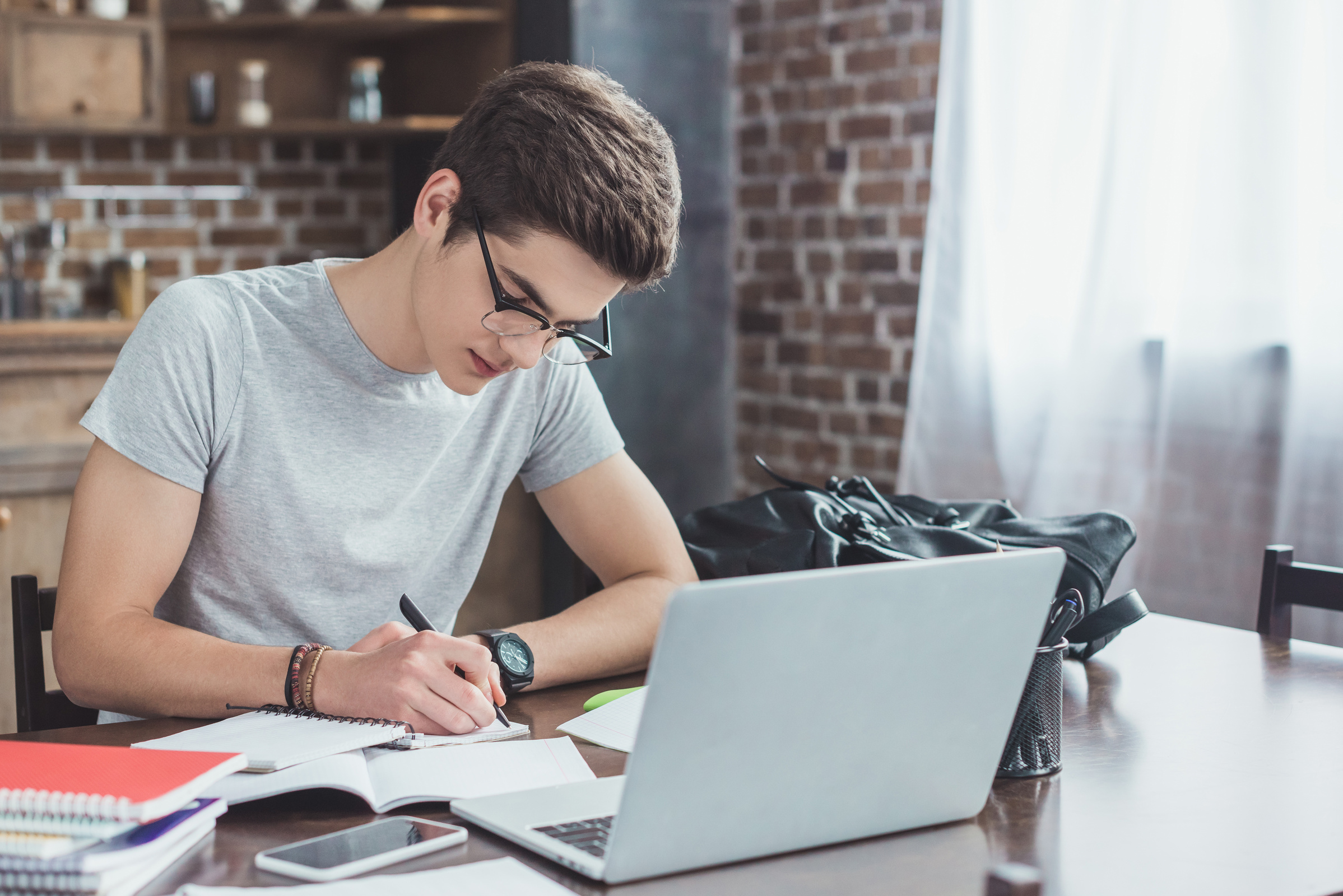Uczeń w okularach siedzi przy biurku i pisze w zeszycie, obok niego znajduje się laptop i smartfon z internetem na kartę