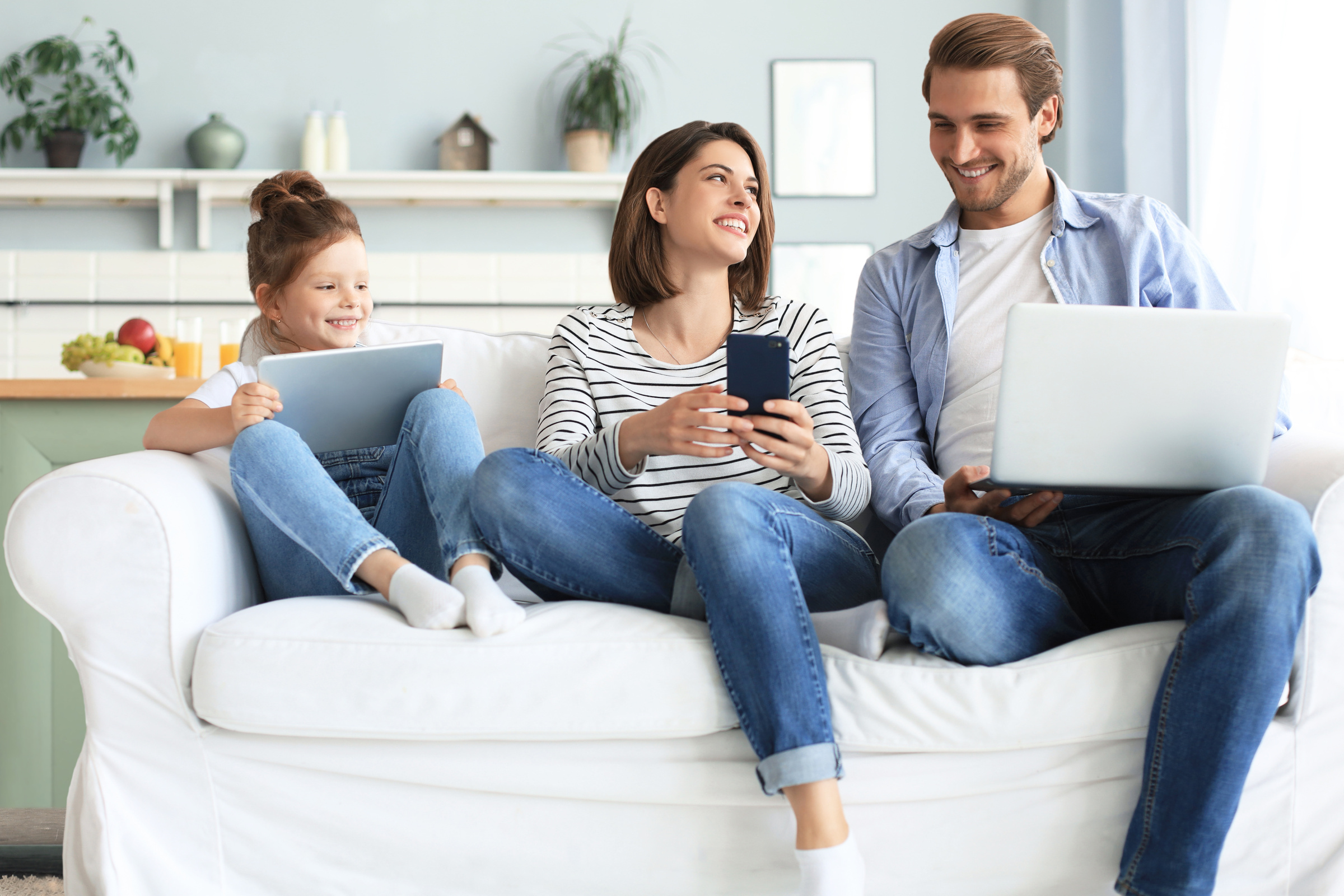 Uśmiechnięta rodzina siedzi razem na kanapie i korzysta z internetu na laptopie, tablecie i smartfonie
