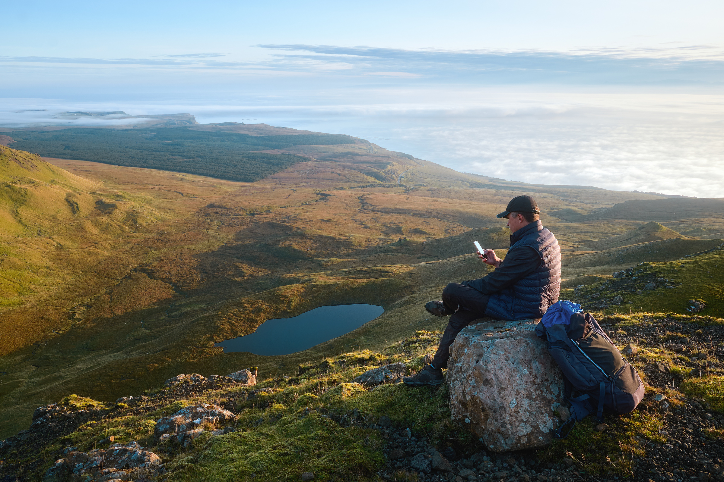 Mężczyzna siedzi na dużej skale w górach, korzysta ze smartfona z internetem na kartę.