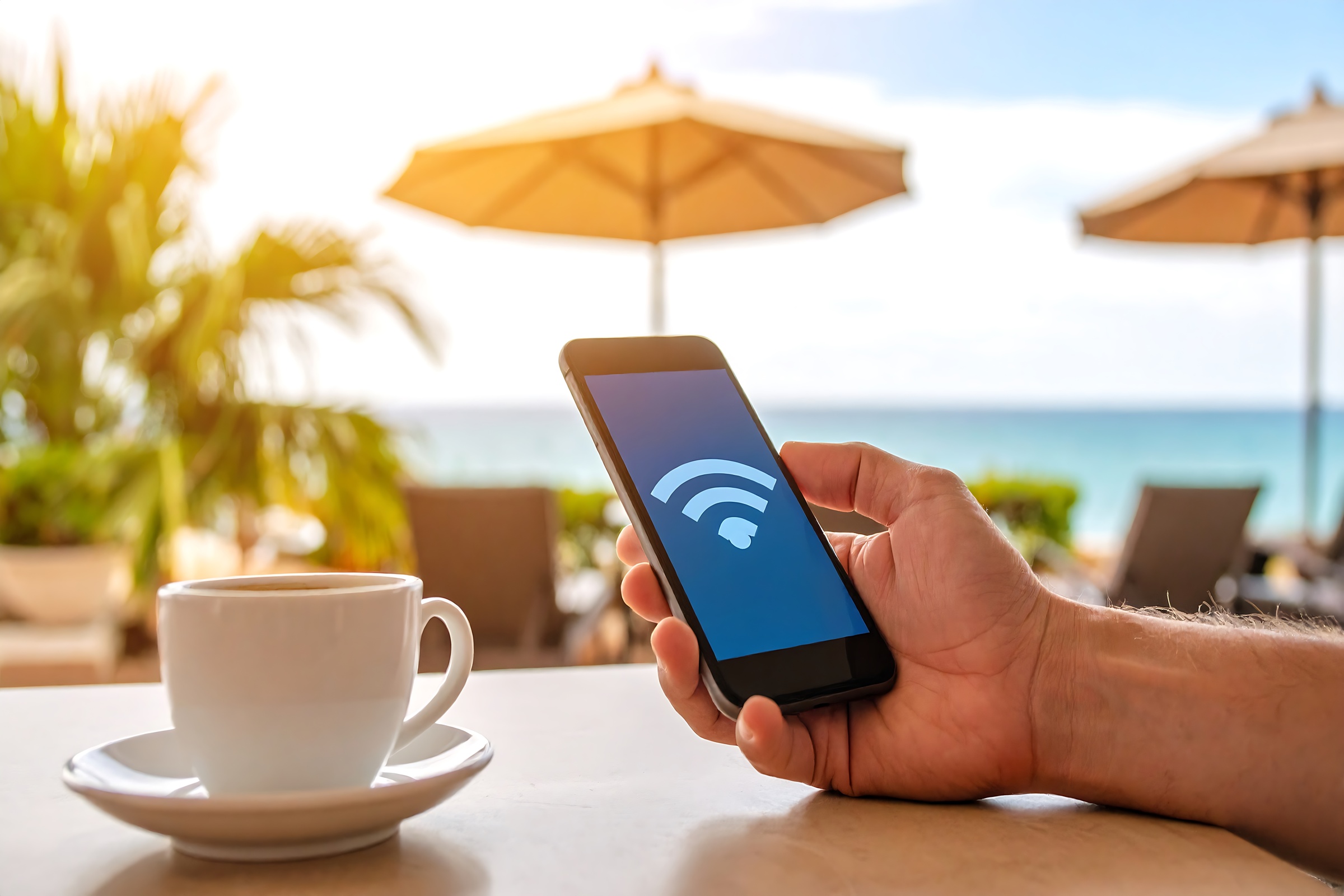 Męska dłoń trzymająca smartfon z niebieskim ekranem i ikoną Wi-Fi, przy stoliku z filiżanką kawy w nadmorskim kurorcie z parasolami w tle.