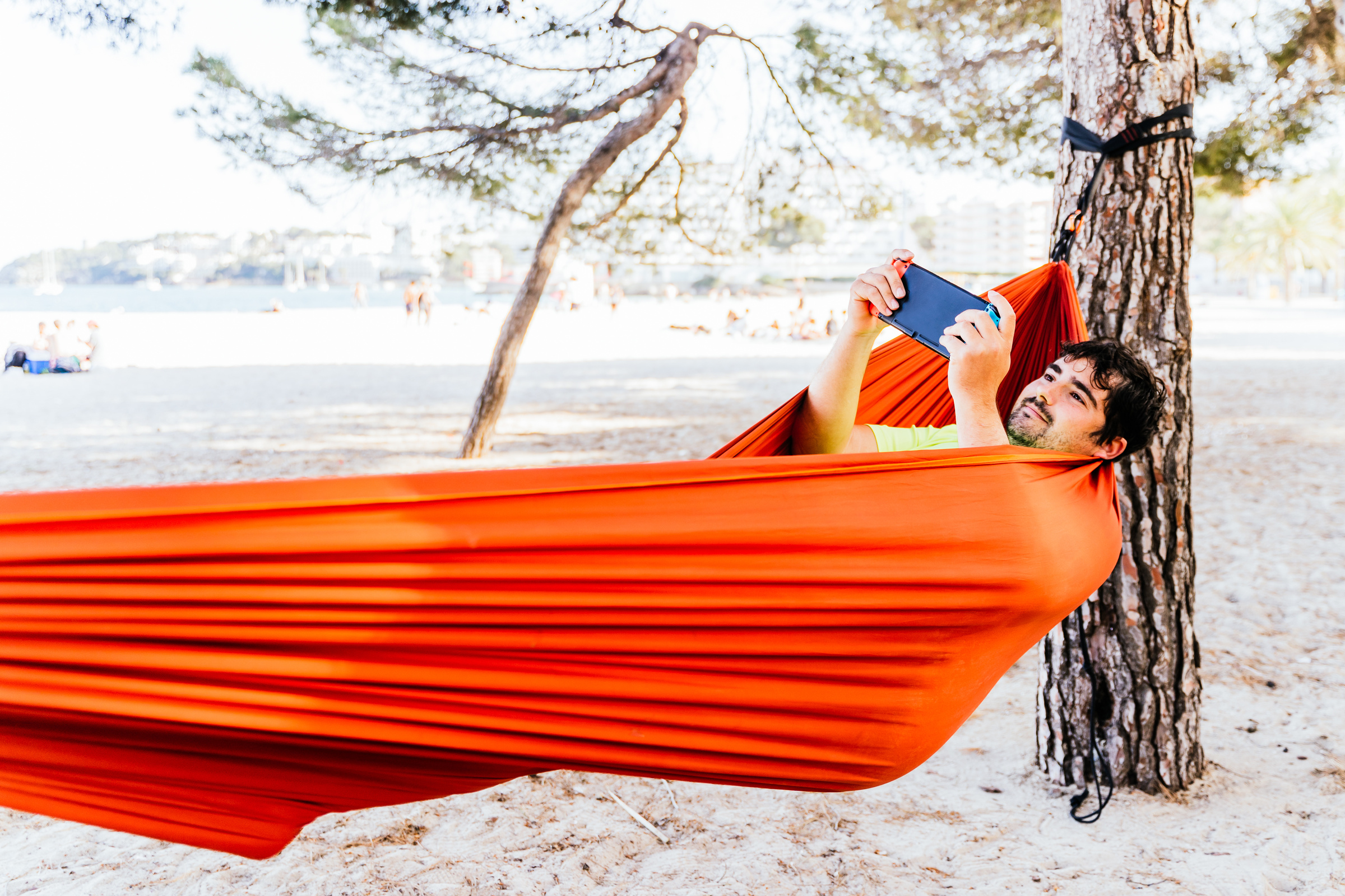 Mężczyzna wypoczywający w pomarańczowym hamaku zawieszonym między drzewami na plaży, trzymający w górze przenośną konsolę do gier.